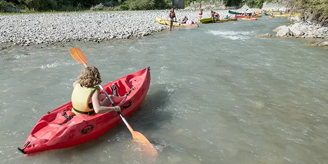 Un enfant en gilet de sauvetage pagaie seul dans un kayak rouge sur la rivière Drôme, rejoint un groupe d’autres enfants et d’adultes sur une plage de galets.