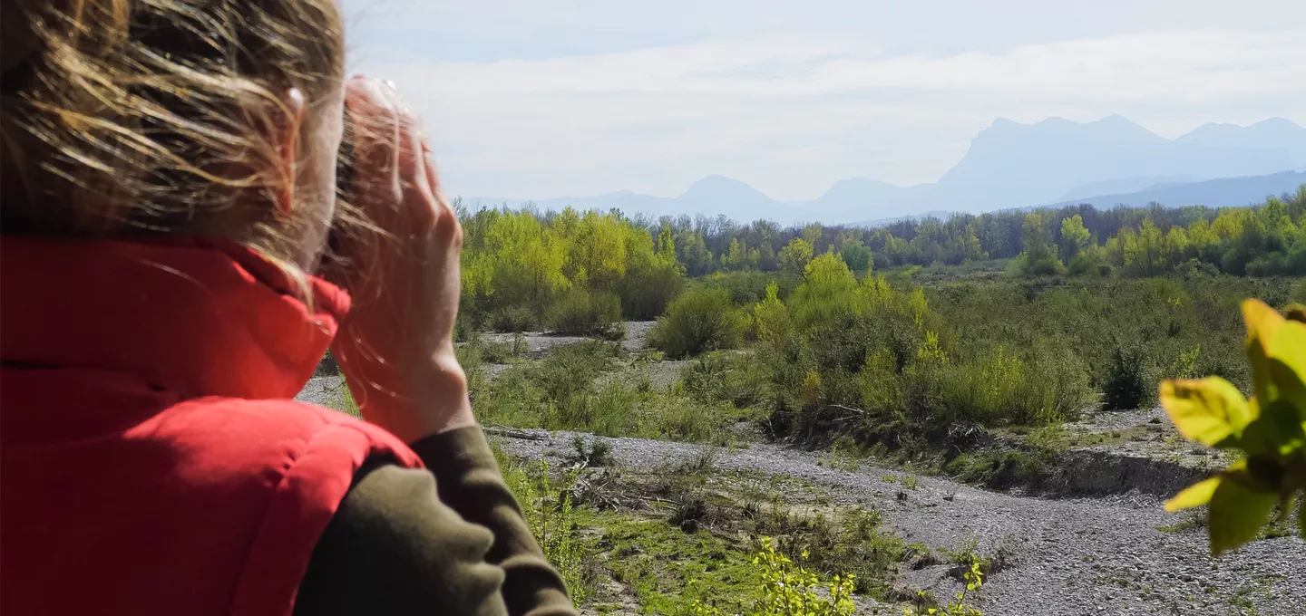 Vue arrière d'une femme en gilet rouge observant la vallée et la rivière Drôme avec des jumelles, face au massif des Trois Becs.
