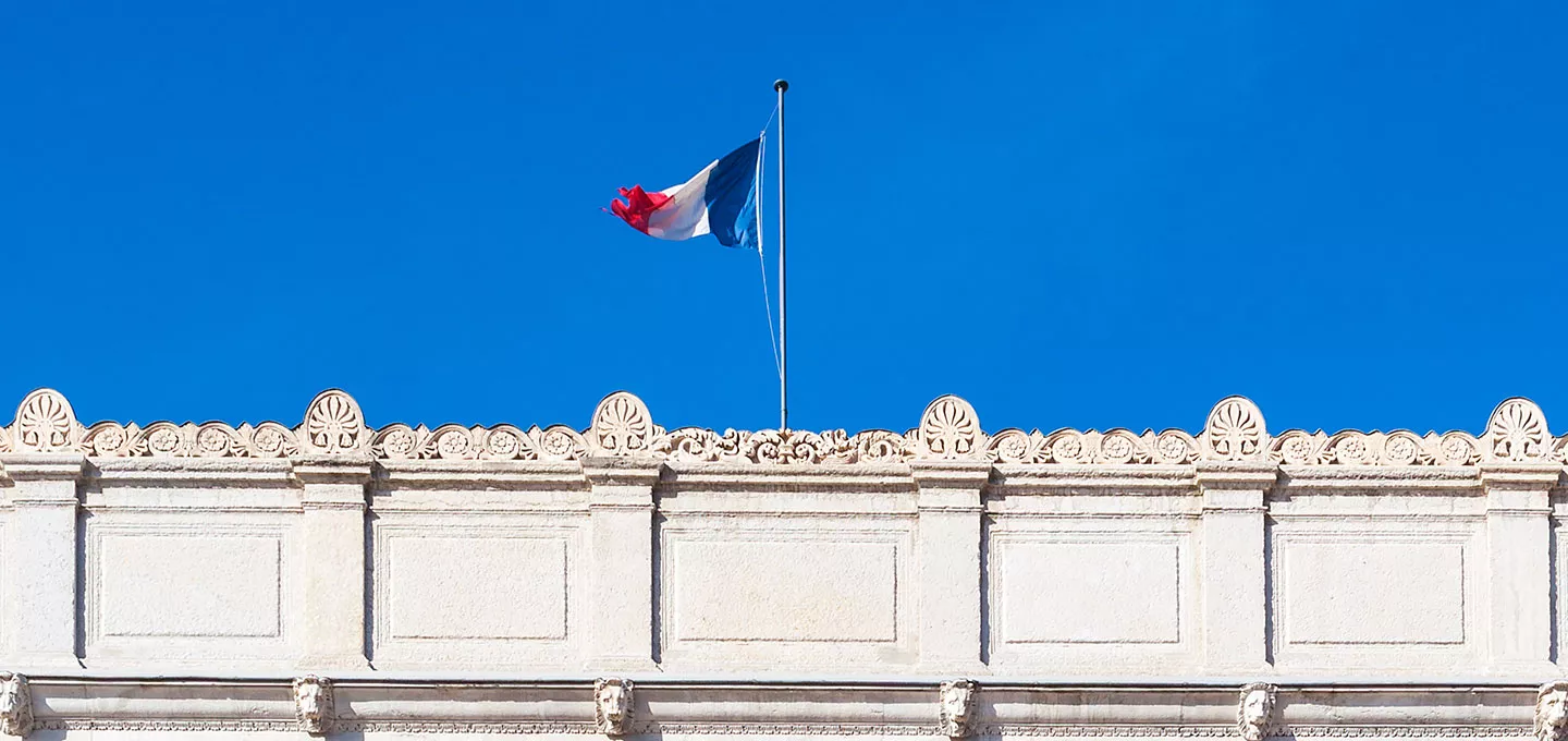 Façade de mairie ornée d’un drapeau tricolore français flottant sur fond de ciel bleu.