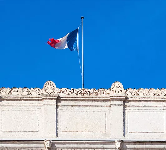 Le drapeau tricolore français flotte au sommet d’un bâtiment public en pierre claire sous un ciel bleu vif.