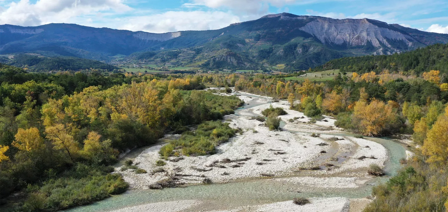 Vue aérienne d’une rivière sinueuse traversant une forêt aux couleurs automnales, avec des montagnes en arrière-plan.