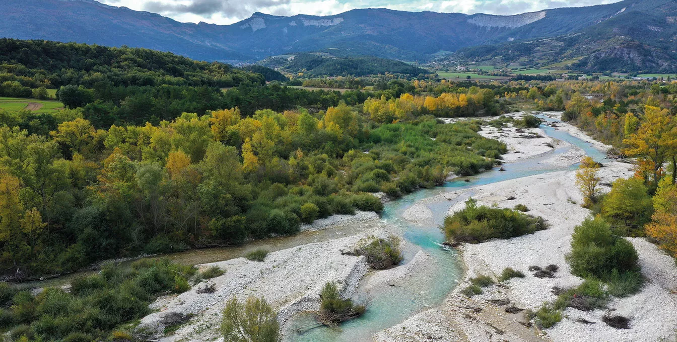 Vue aérienne de la rivière Drôme serpentant au cœur d’une végétation automnale, avec les montagnes en arrière-plan.